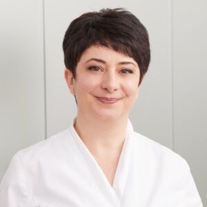 Eine Fachangestellte für podologische Behandlungen im weißen Kittel gekleidet lächelt freundlich in die Kamera. Ihre dunklen Haare trägt sie kurz. Sie ist eine Spezialistin für Spangentechniken und das Modellieren von Orthosen bei der Privatpraxis.