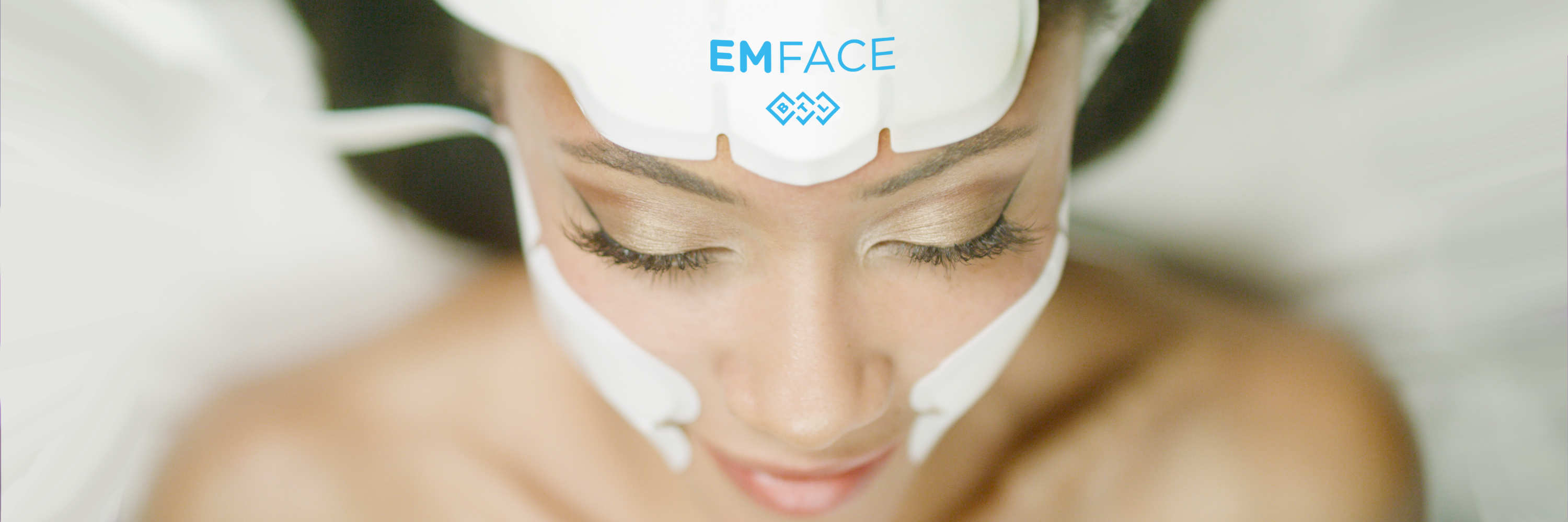 Eine Frau wünscht sich ein Gesicht ohne Falten und einen Lifting-Effekt. Die Abbildung zeigt die liegende Person während einer Faltenbehandlung mit an Stirn und Wangen angeklebten Applikatoren, mit der Aufschrift EMFACE. Sie hat jugendliche Ausstrahlung.